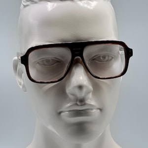 Puede incluir: Gafas con montura marrón de forma cuadrada y lentes transparentes. Las gafas tienen una barra superior y patillas de color marrón oscuro, y se exhiben en una cabeza de maniquí blanca. El estilo general es vintage.