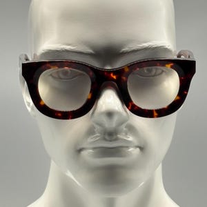 Puede incluir: Gafas con estampado de carey y montura gruesa. Las gafas tienen forma redondeada y se exhiben en una cabeza de maniquí blanca. Las lentes son de color claro.