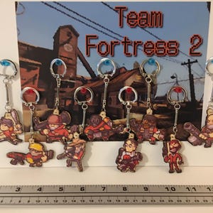 May include: A collection of ten Team Fortress 2 pixel art character keychains featuring various classes, displayed against a game-themed backdrop. A metal ruler at the bottom shows the scale in inches, with the display spanning approximately 13 inches.