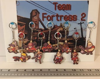 May include: A collection of ten Team Fortress 2 pixel art character keychains featuring various classes, displayed against a game-themed backdrop. A metal ruler at the bottom shows the scale in inches, with the display spanning approximately 13 inches.