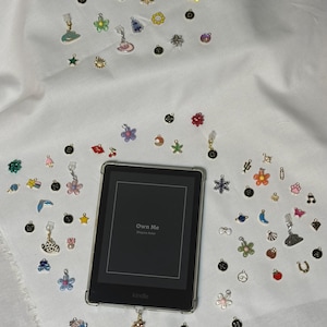 May include: A Kindle e-reader with a clear case, surrounded by various small charms. The charms include flowers, stars, and other decorative shapes. The e-reader displays the text "Own Me" on the screen.