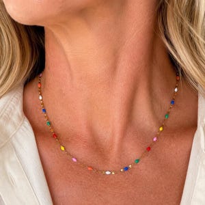 May include: A gold-colored necklace featuring a delicate chain adorned with colorful, oval-shaped beads. The beads are in various colors, including red, yellow, blue, green, and white. The necklace is worn around the neck.