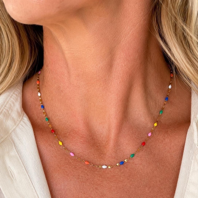 May include: A gold-colored necklace featuring a delicate chain adorned with colorful, oval-shaped beads. The beads are in various colors, including red, yellow, blue, green, and white. The necklace is worn around the neck.