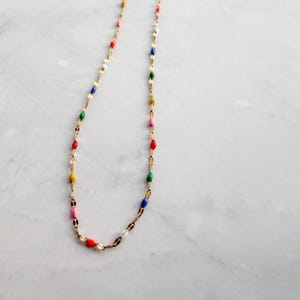 May include: A delicate gold-tone necklace featuring colorful enamel beads in red, yellow, pink, green, blue, and white. The necklace has a fine chain and is displayed on a white marble surface. The beads are evenly spaced along the chain.