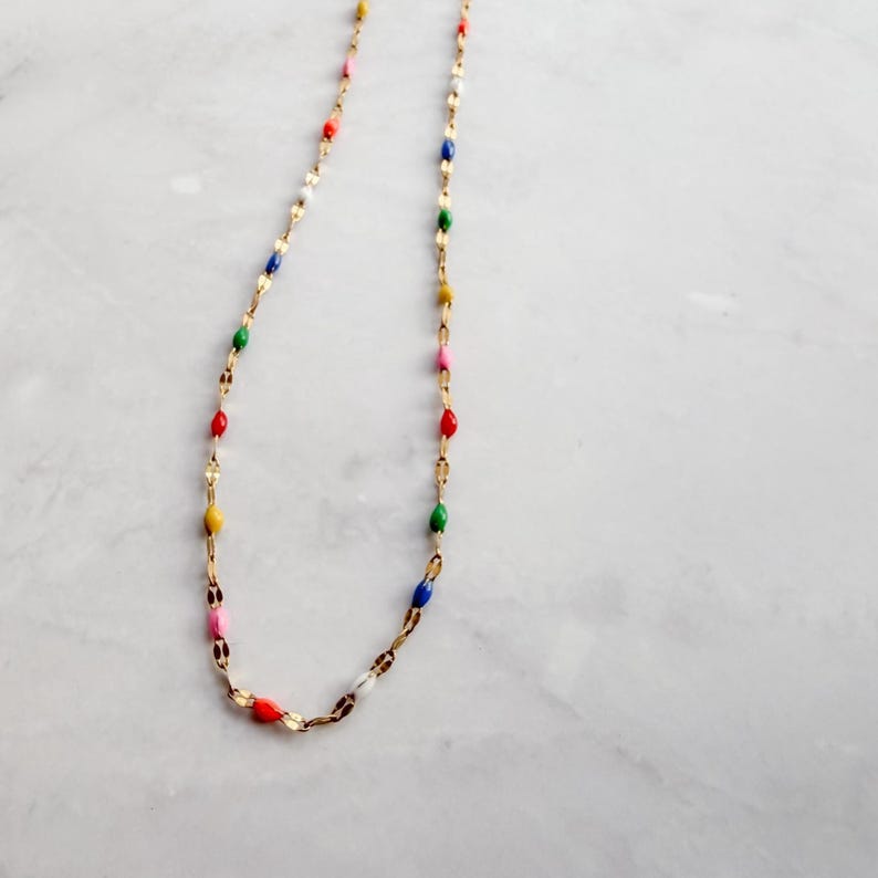 May include: A delicate gold-tone necklace featuring colorful enamel beads in red, yellow, pink, green, blue, and white. The necklace has a fine chain and is displayed on a white marble surface. The beads are evenly spaced along the chain.