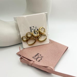 May include: Gold-toned hoop earrings in various sizes are displayed on a white card within a clear plastic sleeve. The card has a logo. A pink pouch with a matching logo is below the earrings.