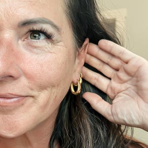 May include: Gold-colored hoop earrings with a textured design. The earrings are worn on the ear of a person. The person's hand is near the ear. The earrings have a shiny finish and a classic, elegant style.