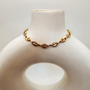 May include: A gold-colored chain necklace with a series of oval-shaped links. The necklace is displayed on a white mannequin bust. The links are connected by smaller, circular connectors, creating a continuous loop. The jewelry is a fashion accessory.