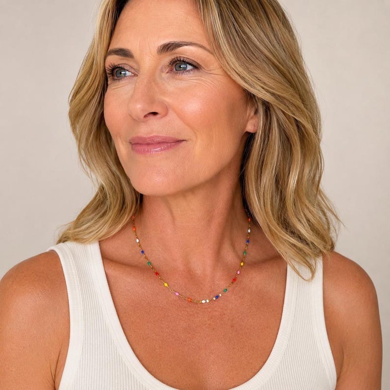 May include: A gold-toned necklace featuring a delicate chain adorned with small, colorful beads. The beads are in various colors, including red, yellow, green, blue, and white. The necklace is worn by a person wearing a white tank top.