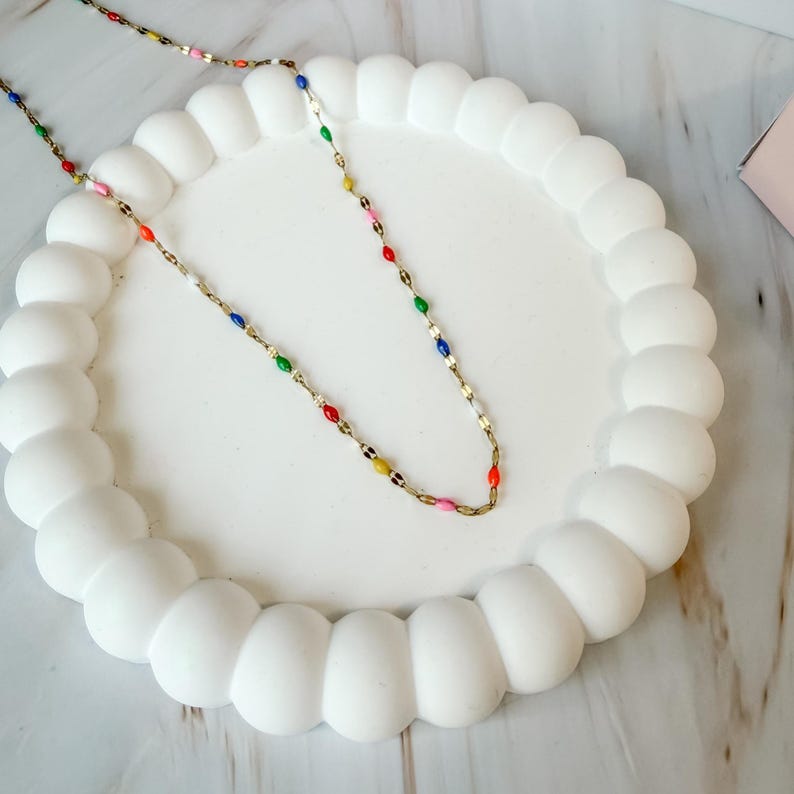 May include: A delicate gold-tone necklace with colorful enamel accents. The necklace features small, oval-shaped beads in red, orange, yellow, green, blue, and pink. It rests on a white, scalloped-edge tray.
