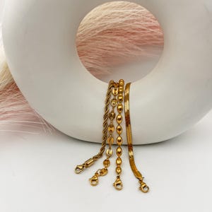 Puede incluir: Cuatro pulseras de cadena doradas con diferentes estilos de eslabones, incluyendo una cadena de cuerda, una cadena de cuentas y una cadena de serpiente. Las pulseras se muestran sobre una superficie de cer&aacute;mica blanca.