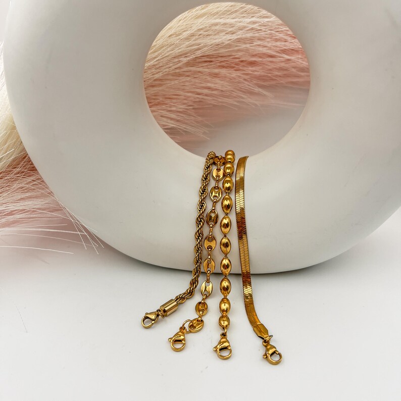 Puede incluir: Cuatro pulseras de cadena doradas con diferentes estilos de eslabones, incluyendo una cadena de cuerda, una cadena de cuentas y una cadena de serpiente. Las pulseras se muestran sobre una superficie de cer&aacute;mica blanca.