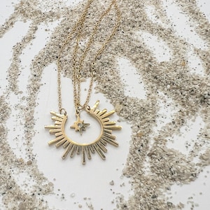 May include: Gold-toned necklace featuring a sunburst pendant and a smaller star pendant with a central gem. The necklace is displayed on a white surface with a scattering of small, light-colored granules.