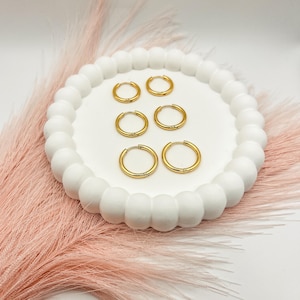 May include: Six gold-colored hoop earrings of varying sizes are arranged on a white, round, beaded tray. The earrings are displayed against a backdrop of pink feathers and a white surface, creating a minimalist aesthetic.