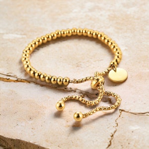 May include: A gold beaded bracelet with a sliding clasp and a small round charm. The bracelet is adjustable and can be worn on the wrist or ankle.