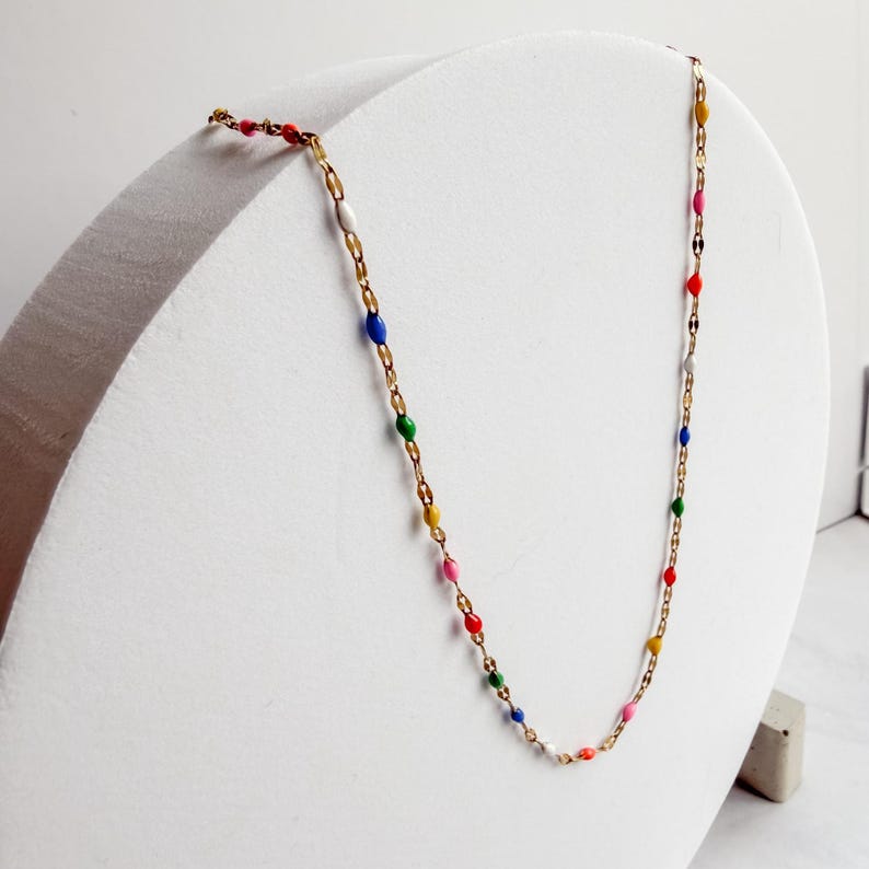 May include: A delicate gold-tone necklace featuring colorful enamel beads. The necklace has a chain design with small, oval-shaped beads in various colors, including red, pink, yellow, green, and blue. The necklace is displayed on a white stand.