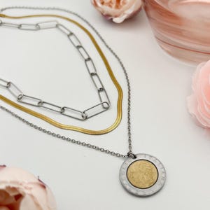 May include: A layered necklace with three chains. The top chain is silver with large rectangular links. The middle chain is gold, and the bottom chain is silver with a round pendant with gold in the center.