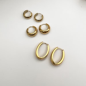 May include: Gold-colored hoop earrings in various sizes. The earrings are arranged on a white surface. There are two pairs of small, round hoops, and one pair of larger, oval-shaped hoops. The earrings have a shiny, polished finish.