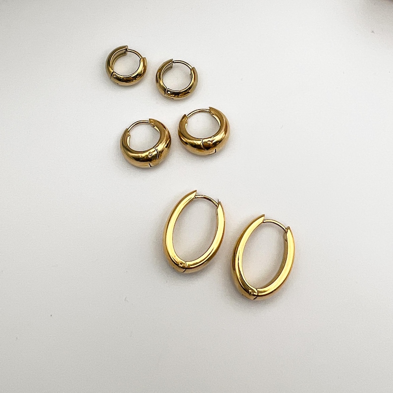May include: Gold-colored hoop earrings in various sizes. The earrings are arranged on a white surface. There are two pairs of small, round hoops, and one pair of larger, oval-shaped hoops. The earrings have a shiny, polished finish.