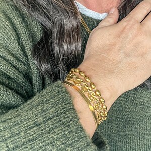 Puede incluir: Una mu&ntilde;eca de mujer con cuatro pulseras de oro. Las pulseras est&aacute;n apiladas una encima de la otra y est&aacute;n hechas de diferentes estilos de cadena. Las pulseras son todas de color dorado.