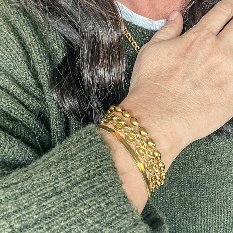 Puede incluir: Una mu&ntilde;eca de mujer con cuatro pulseras de oro. Las pulseras est&aacute;n apiladas una encima de la otra y est&aacute;n hechas de diferentes estilos de cadena. Las pulseras son todas de color dorado.