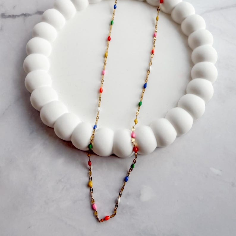 May include: A gold-colored necklace with colorful enamel beads in red, yellow, pink, blue, green, and white. The necklace is displayed on a white, textured, circular tray.
