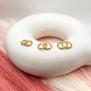 May include: Three pairs of gold hoop earrings of different sizes on a white ceramic surface.