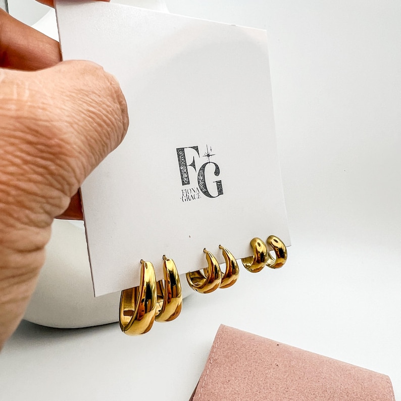 Gold-colored hoop earrings in three different sizes are displayed on a white card with the logo "FG for FIONA GRACE". The earrings are shiny and reflective, with a smooth surface. The earrings are presented on a white background.