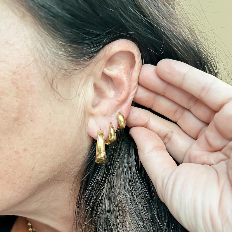 May include: Three gold-colored hoop earrings of varying sizes are worn on an ear. The largest earring is a thick, rounded hoop. The other two are smaller, teardrop-shaped, and also gold. The ear is next to a hand.