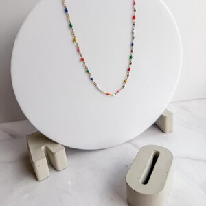May include: A delicate gold-tone necklace with colorful enamel accents. The necklace features small, oval-shaped beads in various colors, including red, blue, green, yellow, and pink. The necklace is displayed on a white circular stand.