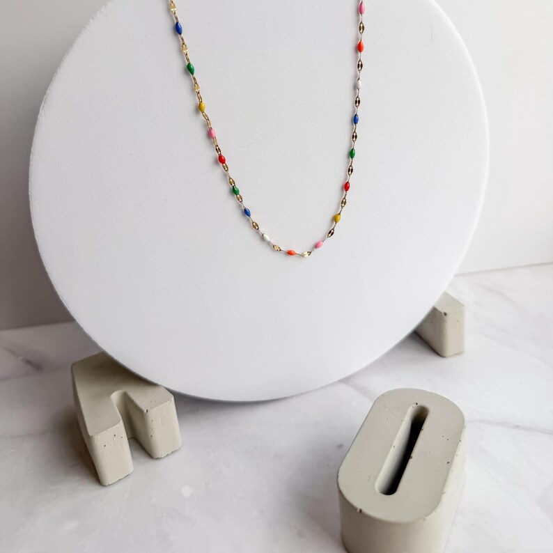 May include: A delicate gold-tone necklace with colorful enamel accents. The necklace features small, oval-shaped beads in various colors, including red, blue, green, yellow, and pink. The necklace is displayed on a white circular stand.
