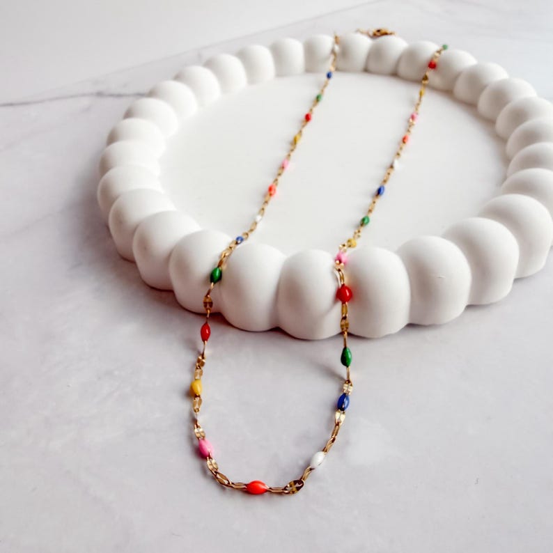 May include: A gold-colored necklace with colorful, oval-shaped beads. The beads are in various colors, including red, green, blue, yellow, and pink. The necklace is displayed on a white, decorative, cloud-shaped tray.
