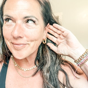 May include: A woman wearing gold hoop earrings and layered gold necklaces with pearl accents. Her wrist displays a stack of colorful beaded bracelets. The woman is smiling and touching her ear with her hand.