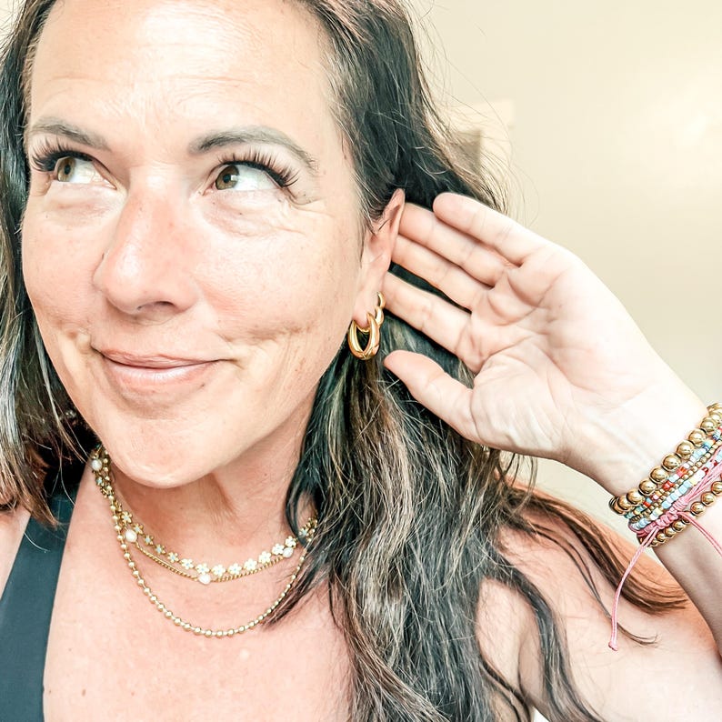 May include: A woman wearing gold hoop earrings and layered gold necklaces with pearl accents. Her wrist displays a stack of colorful beaded bracelets. The woman is smiling and touching her ear with her hand.
