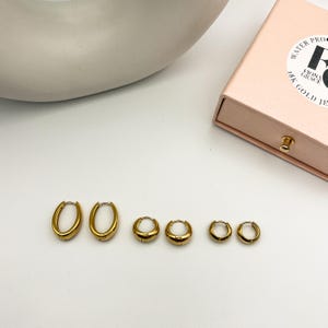 May include: A collection of six gold-colored hoop earrings in varying sizes. The earrings are arranged in a row, showcasing their different dimensions. A pink jewelry box with gold lettering is visible in the background. The earrings appear to be made of a shiny metal.