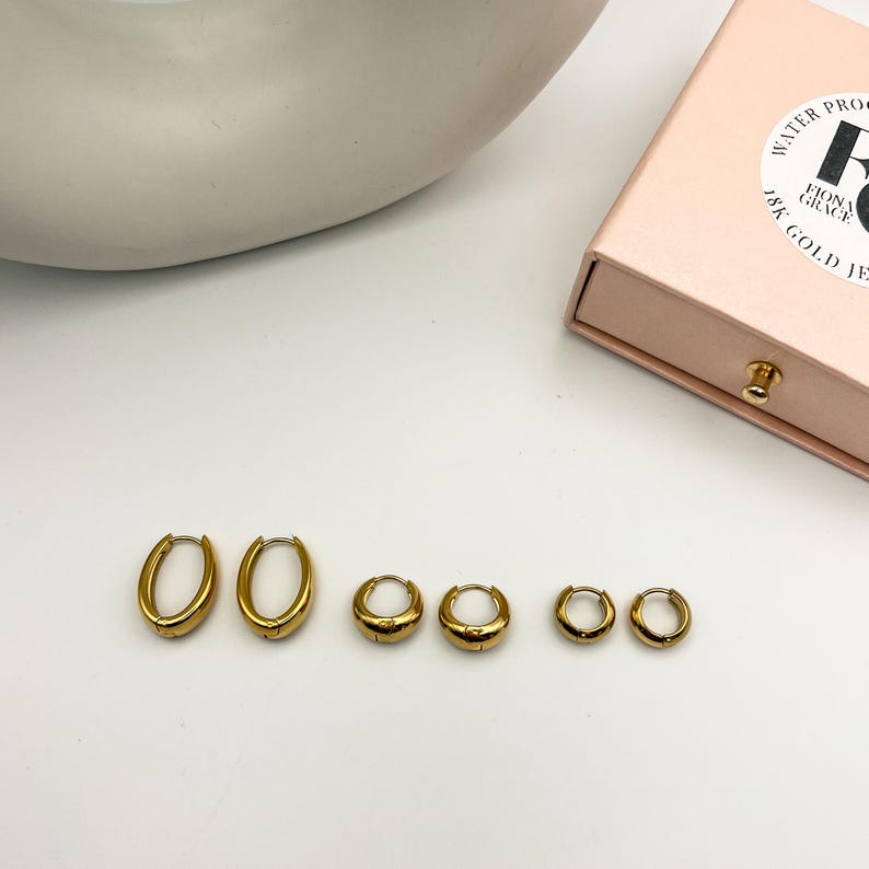 May include: A collection of six gold-colored hoop earrings in varying sizes. The earrings are arranged in a row, showcasing their different dimensions. A pink jewelry box with gold lettering is visible in the background. The earrings appear to be made of a shiny metal.