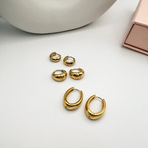 May include: A collection of gold-colored hoop earrings in various sizes. The earrings are displayed on a white surface, with a soft, neutral background. The earrings have a polished finish and a classic design.