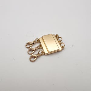 May include: A gold-colored, three-strand necklace clasp with lobster claw clasps. The clasp has a rectangular design with three connecting points for multiple strands. The jewelry component is shown against a white background.