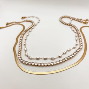 May include: A layered necklace set featuring three gold chains. The top chain has small pearls, the middle chain has clear crystals, and the bottom chain is a flat snake chain. The necklaces are displayed on a white background.