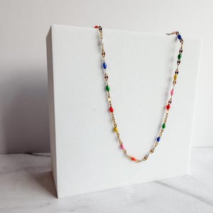 May include: A gold-colored necklace with colorful enamel beads. The necklace features a delicate chain with alternating oval-shaped links and small, colorful beads in red, yellow, pink, green, blue, and white. The necklace is displayed on a white surface.