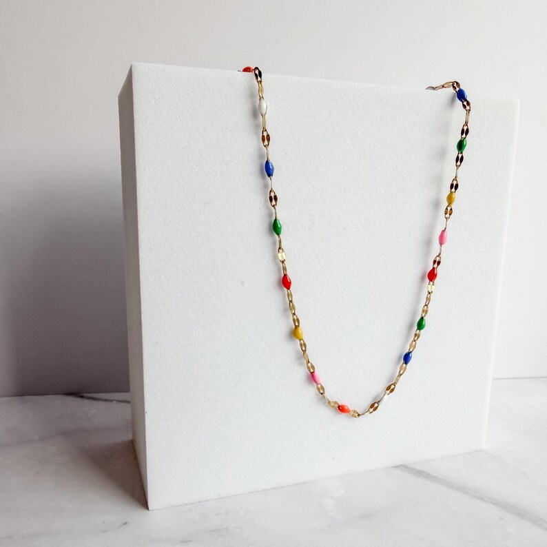 May include: A gold-colored necklace with colorful enamel beads. The necklace features a delicate chain with alternating oval-shaped links and small, colorful beads in red, yellow, pink, green, blue, and white. The necklace is displayed on a white surface.