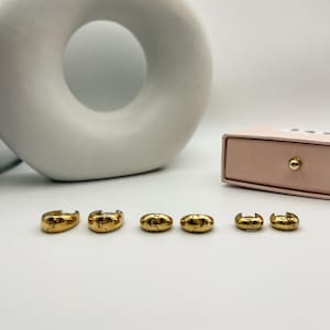 May include: Six gold-colored hoop earrings in various sizes are arranged on a white surface. The earrings have a smooth, polished finish and a simple, classic design. A light pink box with a gold button is in the background.