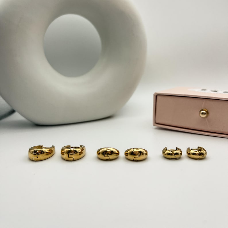 May include: Six gold-colored hoop earrings in various sizes are arranged on a white surface. The earrings have a smooth, polished finish and a simple, classic design. A light pink box with a gold button is in the background.