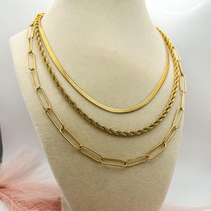 May include: A gold-toned necklace set displayed on a mannequin. The set includes three distinct necklaces: a chain of elongated links, a rope chain, and a flat herringbone chain. The necklaces are layered for a stylish look.