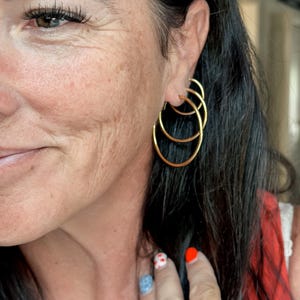 May include: Three stacked gold hoop earrings of varying sizes worn in the earlobe. The earrings are thin, circular, and metallic gold in color, featuring a simple, smooth design that creates a layered appearance when worn together in the ear.