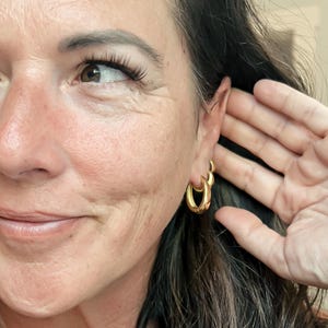 May include: Gold-colored hoop earrings. The earrings are a medium size and have a rounded, polished design. The image shows a close-up of the earrings on a person's ear, with a hand near the ear.