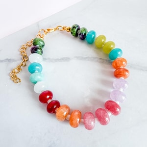 May include: A multi-colored beaded bracelet featuring smooth, round, translucent and opaque gemstone beads in shades of red, orange, yellow, green, blue, and purple. The bracelet includes a gold-toned metal chain extender and a lobster clasp.
