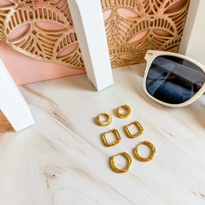 May include: A collection of gold-colored hoop earrings in various shapes, including round and square designs. The earrings are displayed on a marble surface, alongside a pair of white sunglasses.