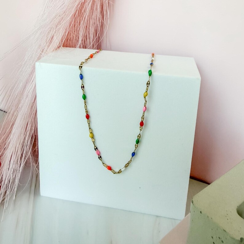 May include: A gold-colored necklace with colorful, oval-shaped beads. The beads are in various colors, including red, yellow, green, blue, pink, and white. The necklace is displayed on a white surface against a pink background.
