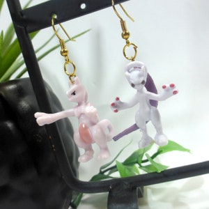 May include: A pair of gold earrings featuring purple and pink Pokemon figures. The figures are of the Pokemon Mewtwo and Mew.